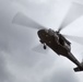 A military helicopter is photographed from below against an overcast sky. The aircraft appears to be a utility/transport helicopter, likely a Sikorsky UH-60 Black Hawk, captured in flight with rotor blades in motion creating a motion blur effect. The image is taken from a low angle looking upward, providing a view of the helicopter's underside.