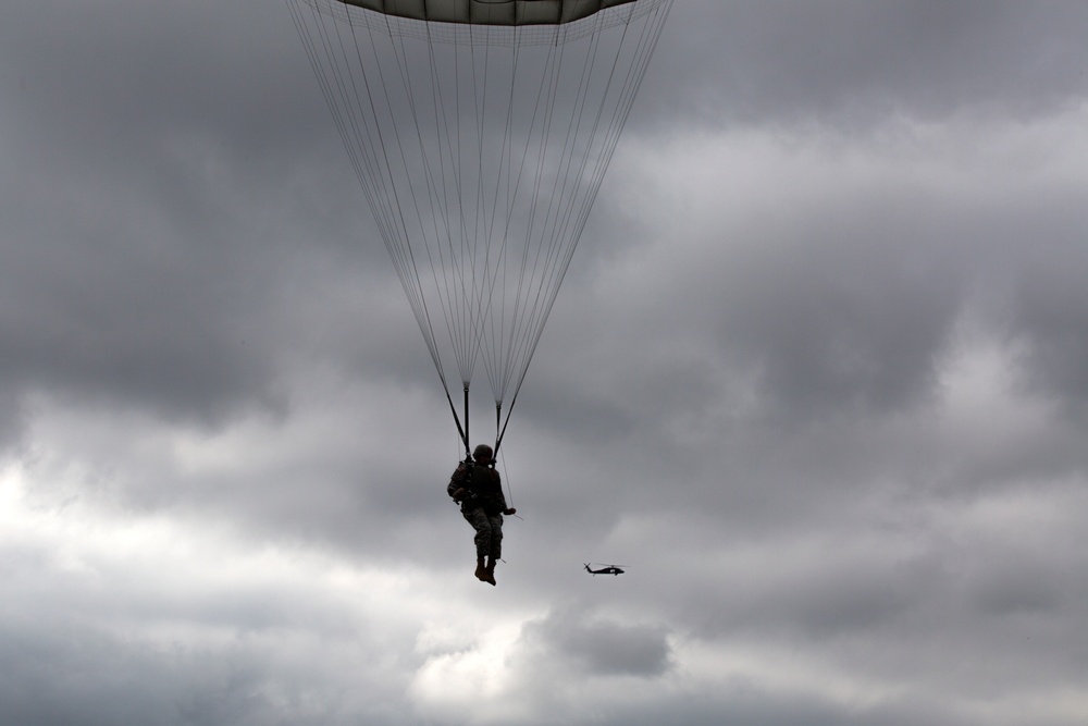 A silhouetted paratrooper descends under a large military-style parachute against a heavily overcast, grey sky. In the background to the right, a helicopter is visible in flight. The image is captured from a low angle looking upward, emphasizing the scale of the parachute canopy.