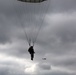 A silhouetted paratrooper descends under a large military-style parachute against a heavily overcast, grey sky. In the background to the right, a helicopter is visible in flight. The image is captured from a low angle looking upward, emphasizing the scale of the parachute canopy.