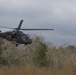 A military helicopter, consistent with a UH-60 Black Hawk, is flying at low altitude over a field of dry grass and sparse woodland. Personnel in military gear are visible seated in the open side door of the aircraft. The overcast sky and bare deciduous trees suggest late autumn or winter conditions.