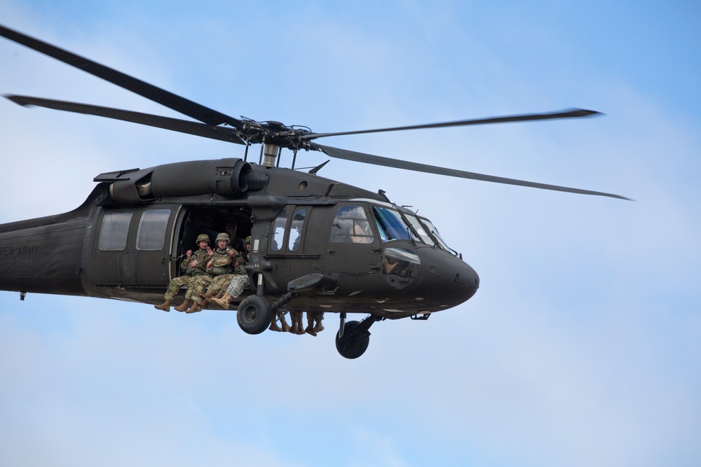 A U.S. Army UH-60 Black Hawk helicopter is captured in flight against a partly cloudy sky. Several soldiers in camouflage uniforms are seated in the open side door of the aircraft with their legs dangling outside. The image is a sharp, close-range aerial photograph showing clear detail of the aircraft and its occupants.