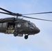 A U.S. Army UH-60 Black Hawk helicopter is captured in flight against a partly cloudy sky. Several soldiers in camouflage uniforms are seated in the open side door of the aircraft with their legs dangling outside. The image is a sharp, close-range aerial photograph showing clear detail of the aircraft and its occupants.