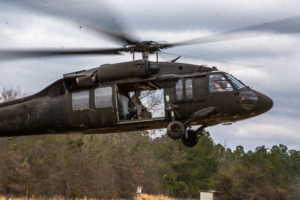 A military UH-60 Black Hawk helicopter is captured in low-altitude flight or hover over a rural, wooded area. The rotor blades are in motion, creating visible debris and downwash in the surrounding air. At least one soldier is visible in the open side door of the aircraft.