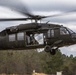 A military UH-60 Black Hawk helicopter is captured in low-altitude flight or hover over a rural, wooded area. The rotor blades are in motion, creating visible debris and downwash in the surrounding air. At least one soldier is visible in the open side door of the aircraft.