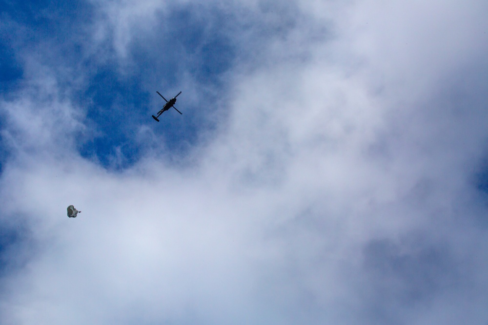 The image shows a helicopter flying at high altitude against a partly cloudy blue sky. Below and to the left of the helicopter, a small parachute with an attached payload or person is visible in descent. The subjects are silhouetted against the sky, suggesting an airdrop or parachuting operation.