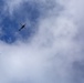 The image shows a helicopter flying at high altitude against a partly cloudy blue sky. Below and to the left of the helicopter, a small parachute with an attached payload or person is visible in descent. The subjects are silhouetted against the sky, suggesting an airdrop or parachuting operation.