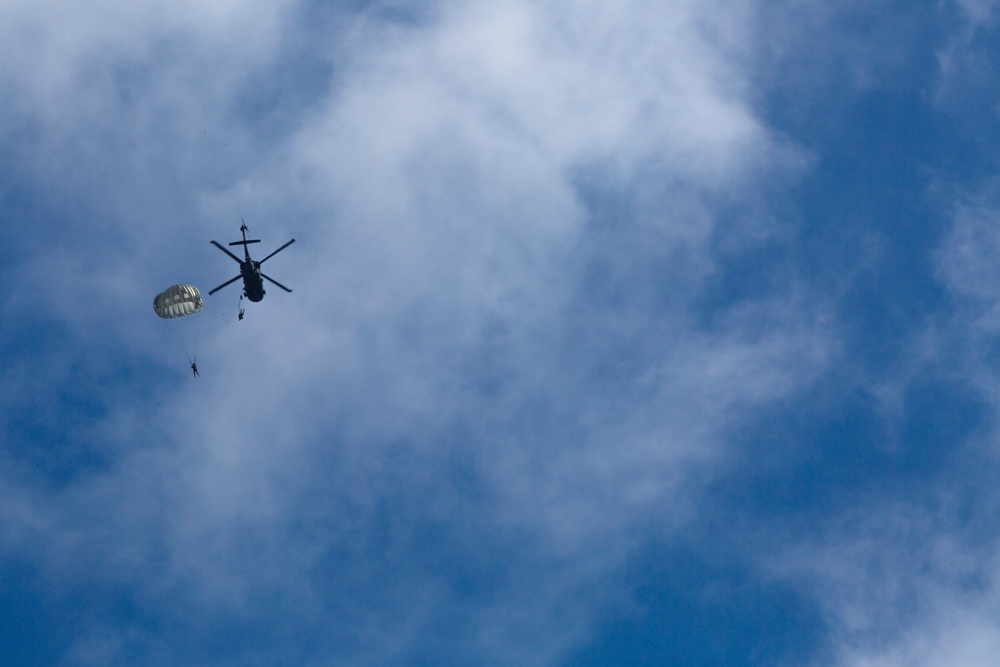 A military helicopter is captured in silhouette against a partly cloudy blue sky, with at least two parachutists visible having jumped from the aircraft. One parachute is fully deployed and visible near the helicopter, while a second figure appears to be in free fall below. This appears to be a military airborne operation or training exercise.