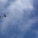 A military helicopter is captured in silhouette against a partly cloudy blue sky, with at least two parachutists visible having jumped from the aircraft. One parachute is fully deployed and visible near the helicopter, while a second figure appears to be in free fall below. This appears to be a military airborne operation or training exercise.