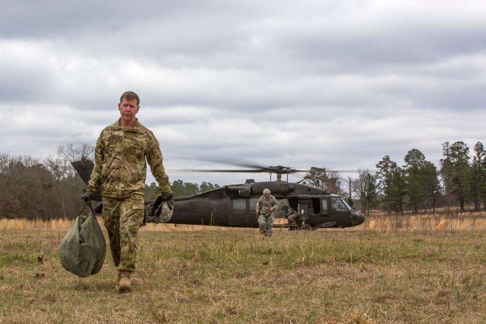 A military soldier in camouflage uniform walks away from a landed UH-60 Black Hawk helicopter in an open field, carrying a large olive-drab bag. Additional personnel are visible near the helicopter, appearing to unload or transfer equipment. The scene suggests a field operation, training exercise, or logistics resupply mission.
