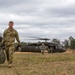 A military soldier in camouflage uniform walks away from a landed UH-60 Black Hawk helicopter in an open field, carrying a large olive-drab bag. Additional personnel are visible near the helicopter, appearing to unload or transfer equipment. The scene suggests a field operation, training exercise, or logistics resupply mission.
