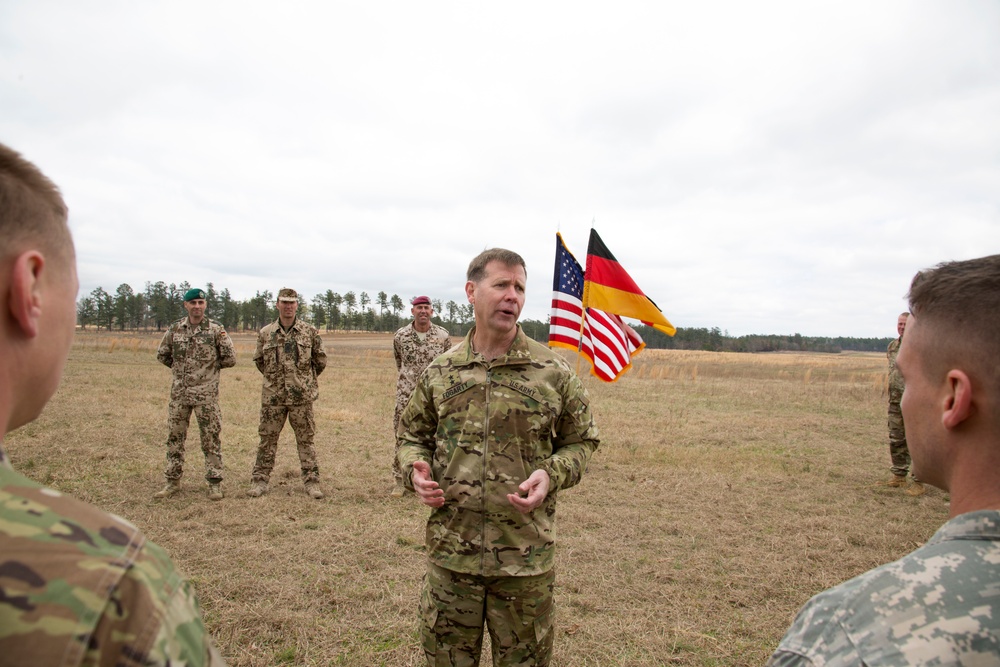 A U.S. Army officer in MultiCam/OCP uniform is addressing a group of soldiers in an open field. Both American and German military personnel are present, as indicated by the different camouflage uniforms and the U.S. and German flags visible in the background. This appears to be a joint military briefing or ceremony between U.S. and German armed forces.