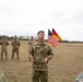 A U.S. Army officer in MultiCam/OCP uniform is addressing a group of soldiers in an open field. Both American and German military personnel are present, as indicated by the different camouflage uniforms and the U.S. and German flags visible in the background. This appears to be a joint military briefing or ceremony between U.S. and German armed forces.