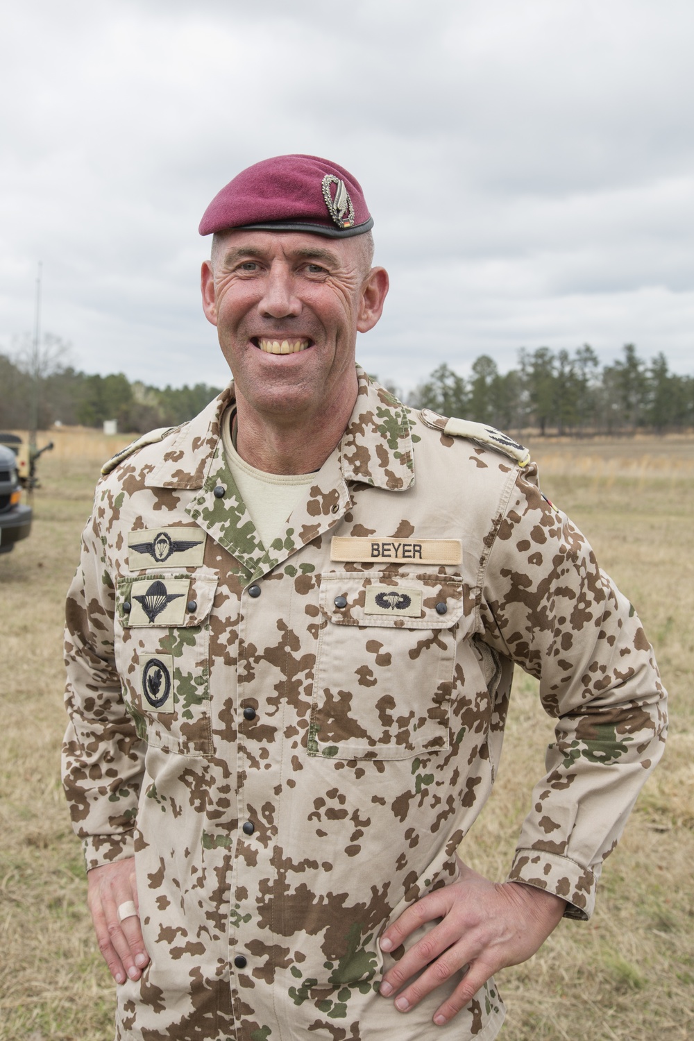 A male military officer wearing German Bundeswehr desert camouflage (Tropentarn) uniform is posing outdoors in a field setting. His name tape reads 'BEYER' and he wears a maroon paratrooper beret with a jump wings badge. He is smiling and standing with hands on hips in what appears to be a training or exercise environment.