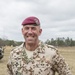 A male military officer wearing German Bundeswehr desert camouflage (Tropentarn) uniform is posing outdoors in a field setting. His name tape reads 'BEYER' and he wears a maroon paratrooper beret with a jump wings badge. He is smiling and standing with hands on hips in what appears to be a training or exercise environment.