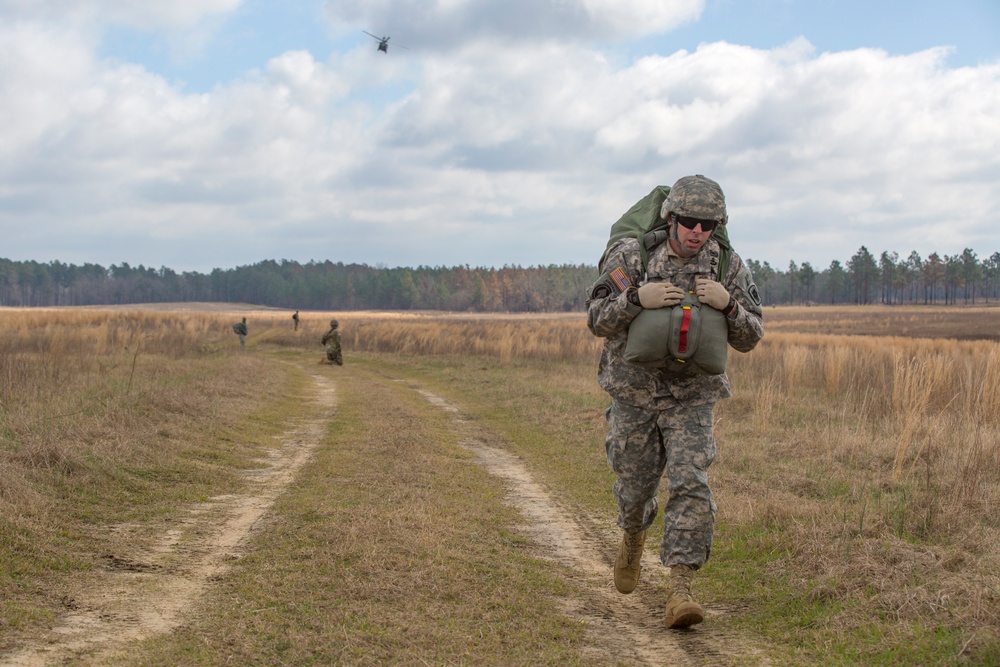 A U.S. Army soldier in ACU (Army Combat Uniform) is running along a dirt trail through an open field while carrying a large rucksack and additional gear. In the background, other soldiers are visible on the field, and a military helicopter (appears to be a Black Hawk) is flying in the distance. The scene appears to be a training exercise or field operation.