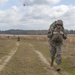 A U.S. Army soldier in ACU (Army Combat Uniform) is running along a dirt trail through an open field while carrying a large rucksack and additional gear. In the background, other soldiers are visible on the field, and a military helicopter (appears to be a Black Hawk) is flying in the distance. The scene appears to be a training exercise or field operation.