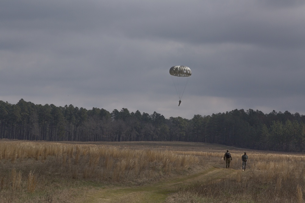 A military parachutist descends toward an open drop zone field under an overcast sky, suspended beneath a round olive-drab parachute canopy. Two individuals on the ground walk along a dirt path across the dry grass field, likely safety or recovery personnel monitoring the landing. A treeline of tall pine trees forms the background horizon.