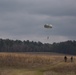A military parachutist descends toward an open drop zone field under an overcast sky, suspended beneath a round olive-drab parachute canopy. Two individuals on the ground walk along a dirt path across the dry grass field, likely safety or recovery personnel monitoring the landing. A treeline of tall pine trees forms the background horizon.
