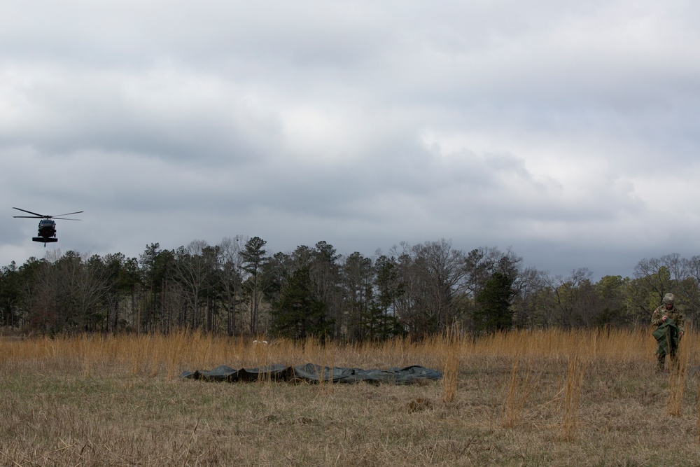 A military helicopter approaches or departs a open field landing zone while a soldier in camouflage uniform stands watch nearby. Dark tarpaulins or equipment bundles are laid out on the dry grass, likely marking or preparing the landing zone. The scene appears to be a military training or logistics operation conducted under overcast winter conditions.