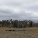 A military helicopter approaches or departs a open field landing zone while a soldier in camouflage uniform stands watch nearby. Dark tarpaulins or equipment bundles are laid out on the dry grass, likely marking or preparing the landing zone. The scene appears to be a military training or logistics operation conducted under overcast winter conditions.