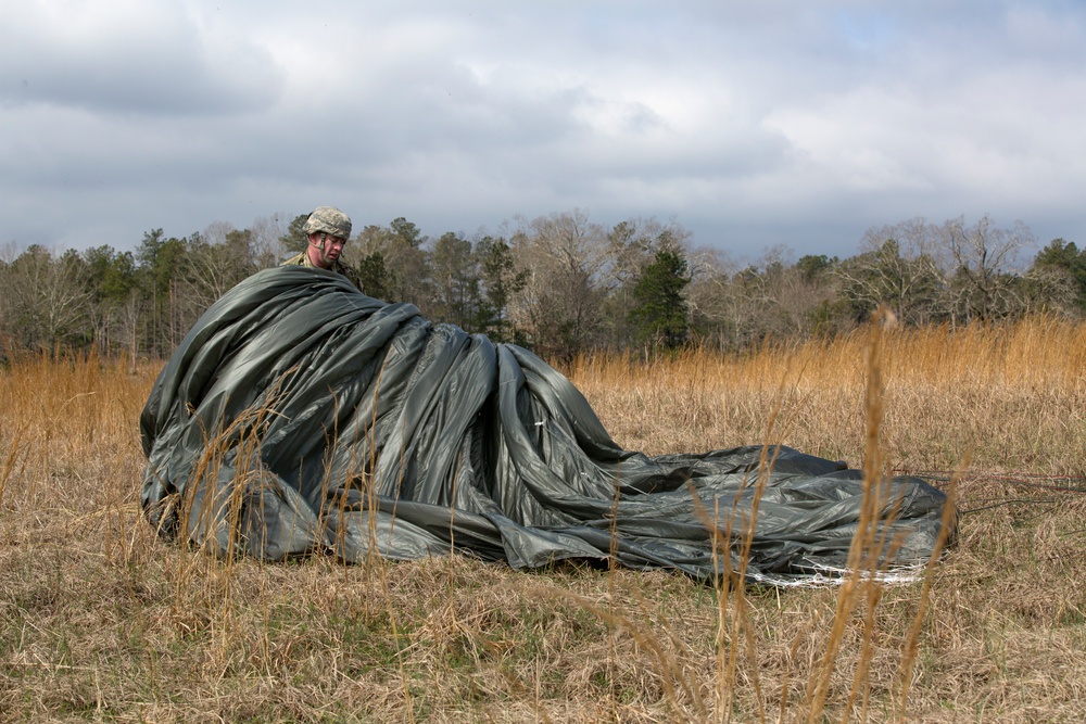 A military paratrooper is seen in an open field gathering and bundling a large olive-drab/dark green parachute canopy after a landing. The soldier is wearing military camouflage uniform and a helmet, and appears to be in the process of recovering and consolidating the parachute. The setting is a dry, grassy field with bare trees in the background under an overcast sky.