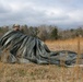 A military paratrooper is seen in an open field gathering and bundling a large olive-drab/dark green parachute canopy after a landing. The soldier is wearing military camouflage uniform and a helmet, and appears to be in the process of recovering and consolidating the parachute. The setting is a dry, grassy field with bare trees in the background under an overcast sky.