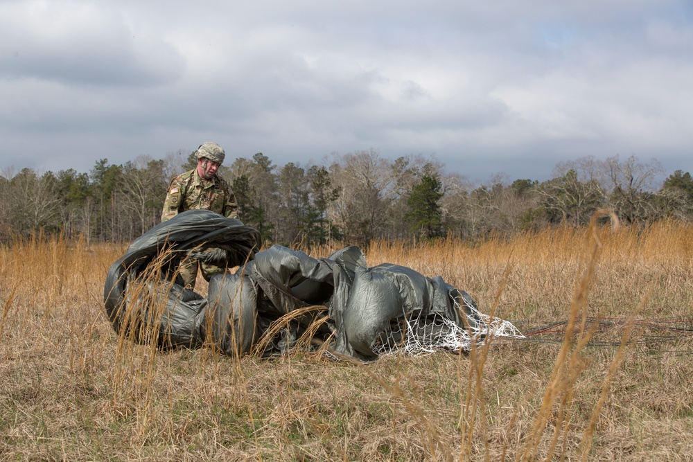 A soldier in camouflage uniform and helmet is gathering or recovering a deployed parachute in an open field with dry, golden grass. The parachute canopy, dark olive/gray in color with visible white netting/suspension lines, is collapsed on the ground. The setting appears to be a military drop zone during late autumn or winter conditions under an overcast sky.