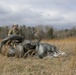 A soldier in camouflage uniform and helmet is gathering or recovering a deployed parachute in an open field with dry, golden grass. The parachute canopy, dark olive/gray in color with visible white netting/suspension lines, is collapsed on the ground. The setting appears to be a military drop zone during late autumn or winter conditions under an overcast sky.