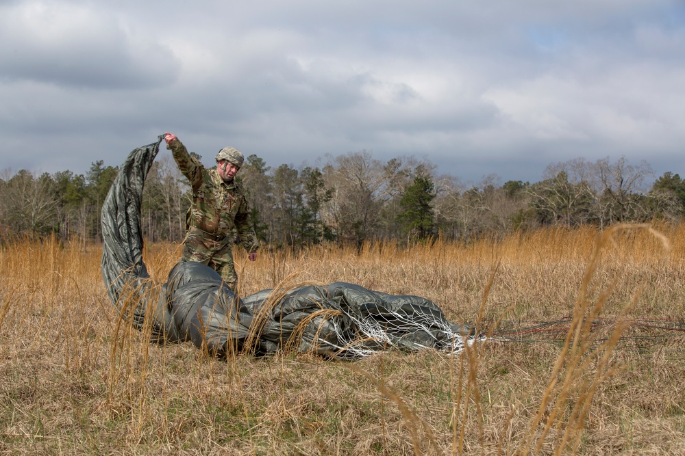 A soldier in camouflage uniform is gathering and recovering a parachute after a landing in an open field. The parachute canopy, which appears to be olive/dark green military-issue, is spread across dry grass on the ground. The setting appears to be a designated drop zone in a rural or training area during overcast conditions.