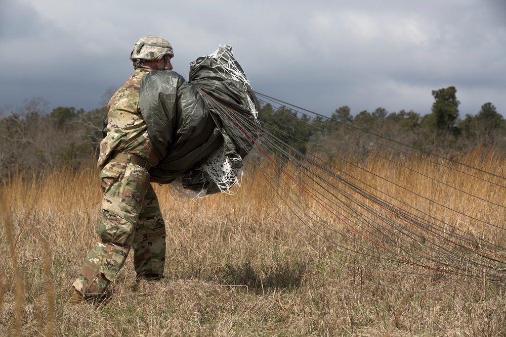 A soldier in full camouflage uniform and helmet is carrying a bundled parachute across an open grassy field after a landing. The parachute canopy and suspension lines trail behind the soldier as they walk away from the camera. The setting appears to be a military drop zone with sparse vegetation and an overcast sky.