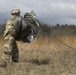 A soldier in full camouflage uniform and helmet is carrying a bundled parachute across an open grassy field after a landing. The parachute canopy and suspension lines trail behind the soldier as they walk away from the camera. The setting appears to be a military drop zone with sparse vegetation and an overcast sky.