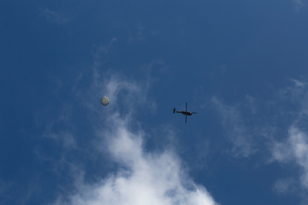 The image shows a helicopter and a deployed parachute captured from a ground-level upward perspective against a blue sky with scattered white clouds. Both objects are at high altitude and appear as silhouettes or small figures due to the distance. The scene suggests a possible airdrop, rescue, or training operation.