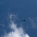 The image shows a helicopter and a deployed parachute captured from a ground-level upward perspective against a blue sky with scattered white clouds. Both objects are at high altitude and appear as silhouettes or small figures due to the distance. The scene suggests a possible airdrop, rescue, or training operation.