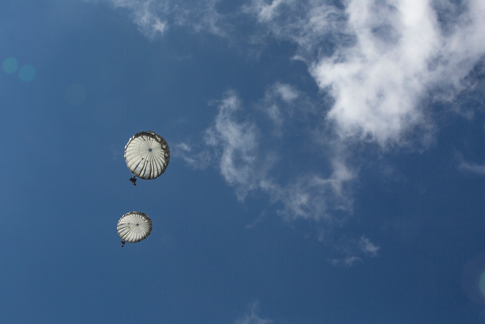 The image shows two parachutists descending against a blue sky with scattered white clouds, photographed from a ground-level upward angle. Both parachutes appear to be round, military-style canopies in a white or light gray color. A lens flare is visible in the upper left corner of the image.
