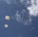 The image shows two parachutists descending against a blue sky with scattered white clouds, photographed from a ground-level upward angle. Both parachutes appear to be round, military-style canopies in a white or light gray color. A lens flare is visible in the upper left corner of the image.