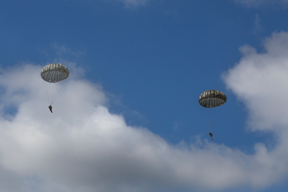 Two military paratroopers are descending through a partly cloudy blue sky using round, olive-drab/gray parachutes. The parachutes appear to be standard military static-line round canopies, consistent with airborne operations. Both jumpers are suspended beneath their fully deployed parachutes at altitude.