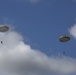 Two military paratroopers are descending through a partly cloudy blue sky using round, olive-drab/gray parachutes. The parachutes appear to be standard military static-line round canopies, consistent with airborne operations. Both jumpers are suspended beneath their fully deployed parachutes at altitude.