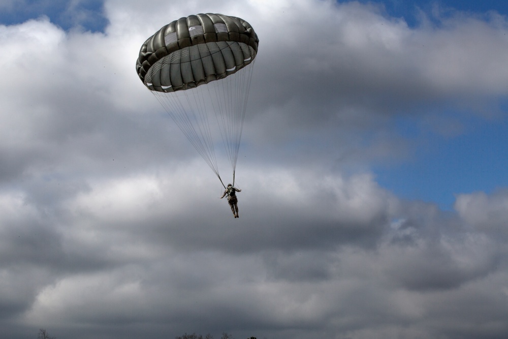 A single parachutist descends through a partly cloudy sky using a round, olive-drab military-style parachute. The jumper appears to be in full gear and is suspended beneath the canopy by multiple suspension lines. The image is taken from a low angle looking upward, with a mix of blue sky and dense cloud cover in the background.
