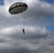 A single parachutist descends through a partly cloudy sky using a round, olive-drab military-style parachute. The jumper appears to be in full gear and is suspended beneath the canopy by multiple suspension lines. The image is taken from a low angle looking upward, with a mix of blue sky and dense cloud cover in the background.