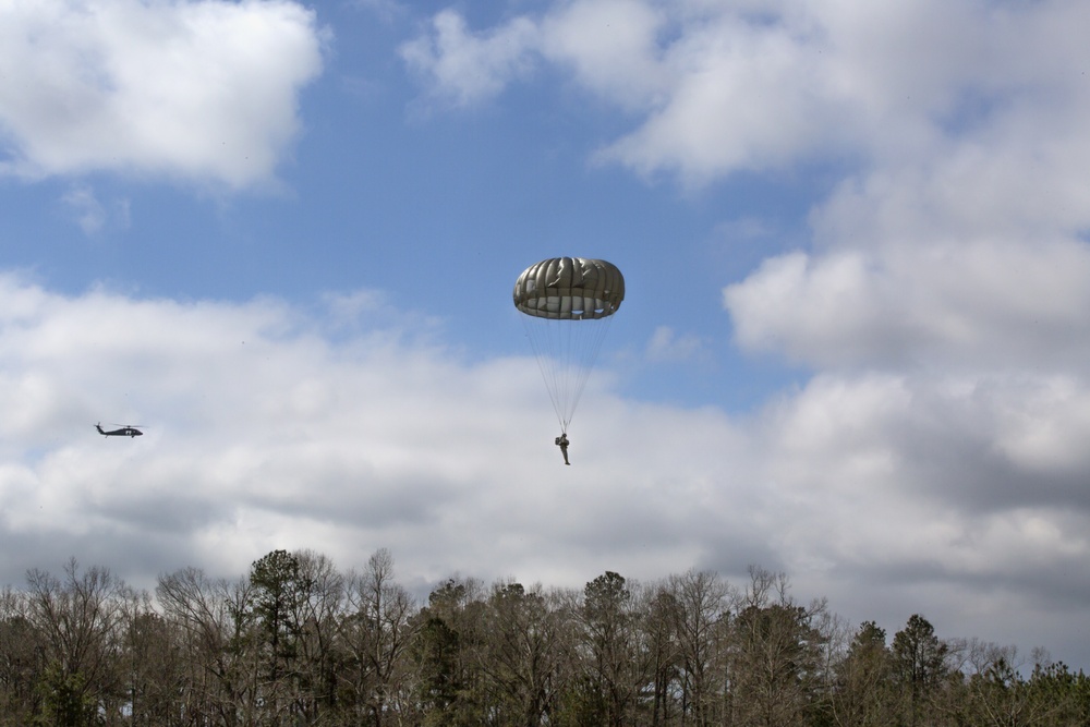 A military paratrooper descends under a deployed olive-drab round parachute over a wooded area. A military helicopter, appearing to be a UH-60 Black Hawk, is visible in the background to the left. The scene appears to be a training or operational airborne jump exercise conducted in a rural, forested environment.