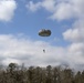 A military paratrooper descends under a deployed olive-drab round parachute over a wooded area. A military helicopter, appearing to be a UH-60 Black Hawk, is visible in the background to the left. The scene appears to be a training or operational airborne jump exercise conducted in a rural, forested environment.