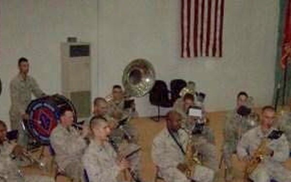 A military band composed of personnel in camouflage uniforms is rehearsing or performing in an indoor facility. The musicians are seated in a semicircular arrangement facing a conductor standing with his back to the camera. American flags and military unit colors are displayed on the walls in the background.