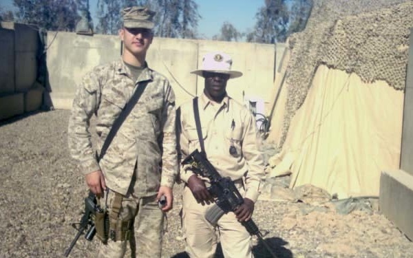 Two armed personnel are posing for a photo at what appears to be a forward operating base or military compound. One individual wears digital camouflage uniform with a patrol cap, while the other wears tan/khaki clothing with a wide-brimmed hat and a badge, suggesting a local security or police role. Both are carrying assault rifles and the background shows concrete barriers, camouflage netting, and gravel terrain.