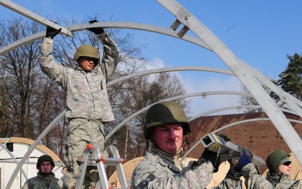 Military personnel in US Army/Air Force digital camouflage uniforms (ACU/ABU pattern) are constructing a large arched metal frame structure, likely a military tent or shelter system. Several soldiers are working collaboratively, with one standing on a Little Giant ladder system to connect overhead frame components. The setting appears to be a military training or deployment area with existing tent structures visible in the background.