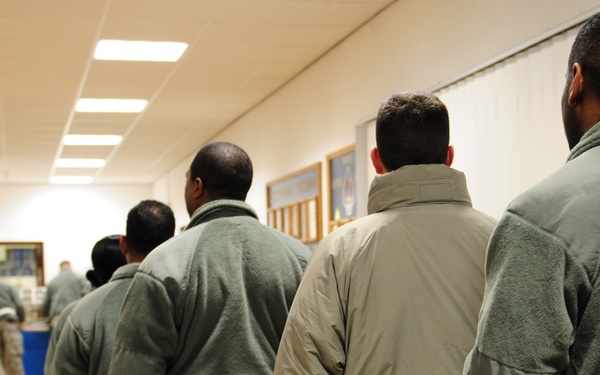 Military personnel in uniform are standing in a chow line inside what appears to be a military dining facility (DFAC). The individuals are wearing various cold-weather military garments including fleece jackets and insulated outerwear. In the background, other service members are seated at tables eating their meals.