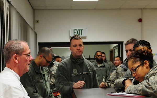 A civilian in a white chef's coat sits at a wooden desk with an open red cash drawer, interacting with a group of military personnel in camouflage uniforms. A female service member wearing glasses and a neon yellow reflective band is writing on a clipboard or document at the desk. The setting appears to be an administrative or check-in area inside a military facility.