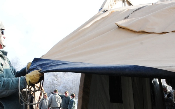 Military personnel in US Air Force uniforms are setting up or dismantling a large military field tent in cold winter conditions. The primary subject in the foreground is pulling on a dark canvas flap of the tent structure while wearing cold-weather gear including a helmet and gloves. Several other service members are visible in the background observing or assisting.