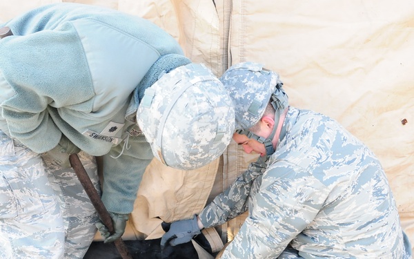 Two military personnel in US Air Force digital camouflage (ABU - Airman Battle Uniform) are working together to secure or stake down the base of a tan military tent. They are working on gravel ground in what appears to be cold weather conditions, as evidenced by their fleece cold-weather gear layered over their uniforms. The activity appears to be tent setup or maintenance at a field installation.