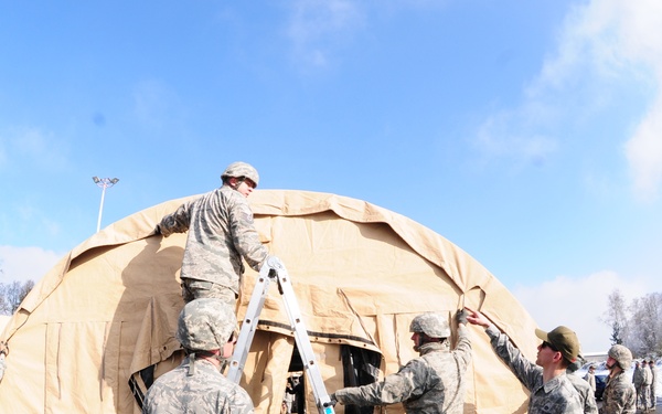 A group of military personnel in US Air Force Airman Battle Uniform (ABU) camouflage pattern are assembling a large tan/beige military field tent in a paved outdoor area during winter conditions. One service member is elevated on a ladder working on the top of the dome-shaped tent while others assist from ground level. The scene appears to be a field training exercise or operational deployment setup.