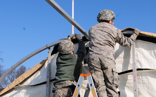 Military personnel in camouflage uniforms are working together to erect or repair a large tent structure. Two individuals are climbing an orange aluminum ladder to work on the upper portion of the tent, while others on the ground provide support and assistance. The scene appears to be an outdoor military field exercise or base camp setup under clear blue skies.